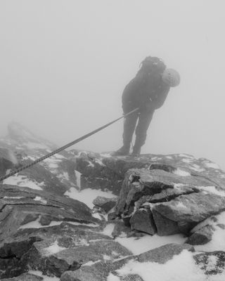 Photo en noir et blanc : Paysage de neige, rochers, brouillard, un alpiniste au rappel cherche la voie