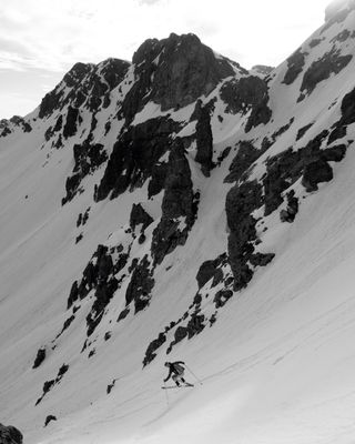 Photo en noir et blanc : Paysage de neige, rochers, un skieur en contrebas effectue un virage sauté