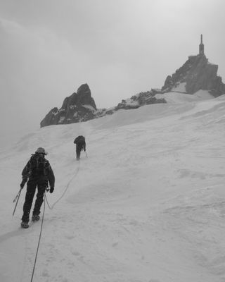 Photo en noir et blanc : Paysage de neige, météo maussade, deux alpinistes remontent vers l'Aiguille du Midi