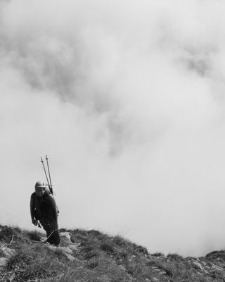 Photo en noir et blanc : une randonneuse encordée, bâtons sur le sac tels deux antennes, remonte une épaule herbeuse. Arrière-plan très nuageux