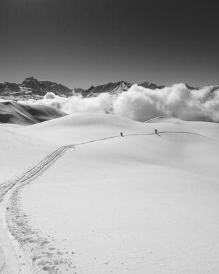 Photo en noir et blanc : Paysage de neige, chaîne de montagnes, nuages, deux skieurs au loin suivent la trace de montée